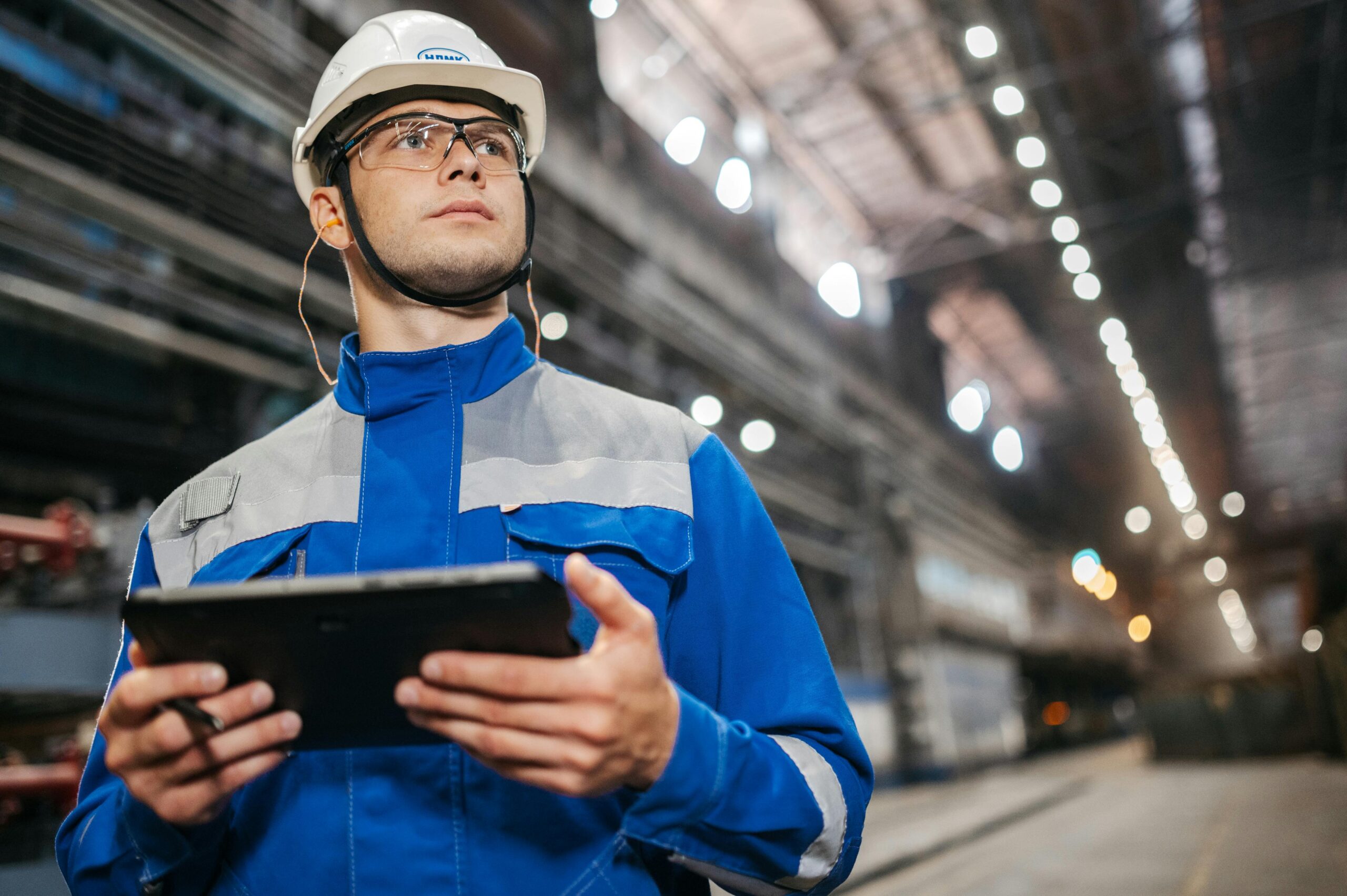 Homme ingénieur ou technicien en casque et veste de travail bleue dans une usine, tenant une tablette, dans une ambiance industrielle. Stratégie digital pour BTP intégrée subtilement.